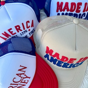 May include: A collection of trucker hats in red, white, and blue. The hats feature text such as "AMERICA", "MADE IN AMERICA", and "AMERICAN HONEY". The hats have mesh backs and curved brims.