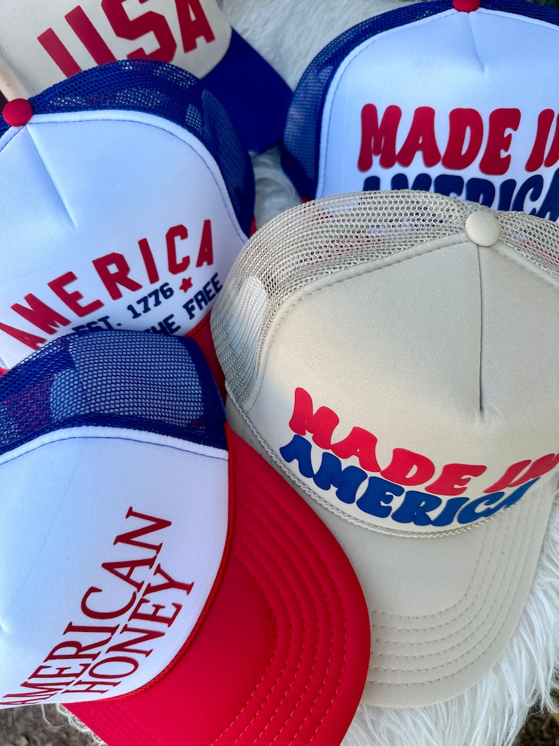 May include: A collection of trucker hats in red, white, and blue. The hats feature text such as "AMERICA", "MADE IN AMERICA", and "AMERICAN HONEY". The hats have mesh backs and curved brims.