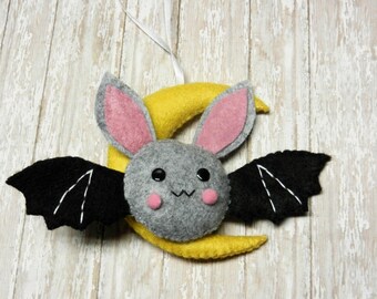 May include: A gray felt bat with pink ears and black wings is hanging from a yellow felt crescent moon. The bat has black eyes and pink cheeks.