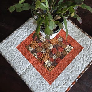 May include: A square quilted table topper featuring a central panel with a dark brown floral pattern in shades of orange, cream, and brown. It is bordered by a solid orange frame with swirling stitch patterns and a light cream outer border.