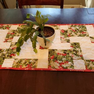 May include: A rectangular quilted table runner featuring a patchwork design of solid white fabric blocks and a festive green holly and red berry print. The runner is finished with a thin red border and a white ceramic pot containing a green plant.