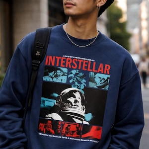 May include: A navy blue crewneck sweatshirt featuring a graphic print with the word INTERSTELLAR in red text. The design includes a central illustration of an astronaut in a helmet, surrounded by smaller panels with blue and red color tones.