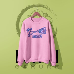 May include: A light pink long-sleeve crewneck sweatshirt featuring a blue graphic print of a revolver and the text The BANG BANG BAR. The design mimics a neon sign style with outlined lettering and a gun silhouette centered on the chest area.