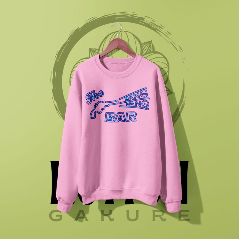 May include: A light pink long-sleeve crewneck sweatshirt featuring a blue graphic print of a revolver and the text The BANG BANG BAR. The design mimics a neon sign style with outlined lettering and a gun silhouette centered on the chest area.
