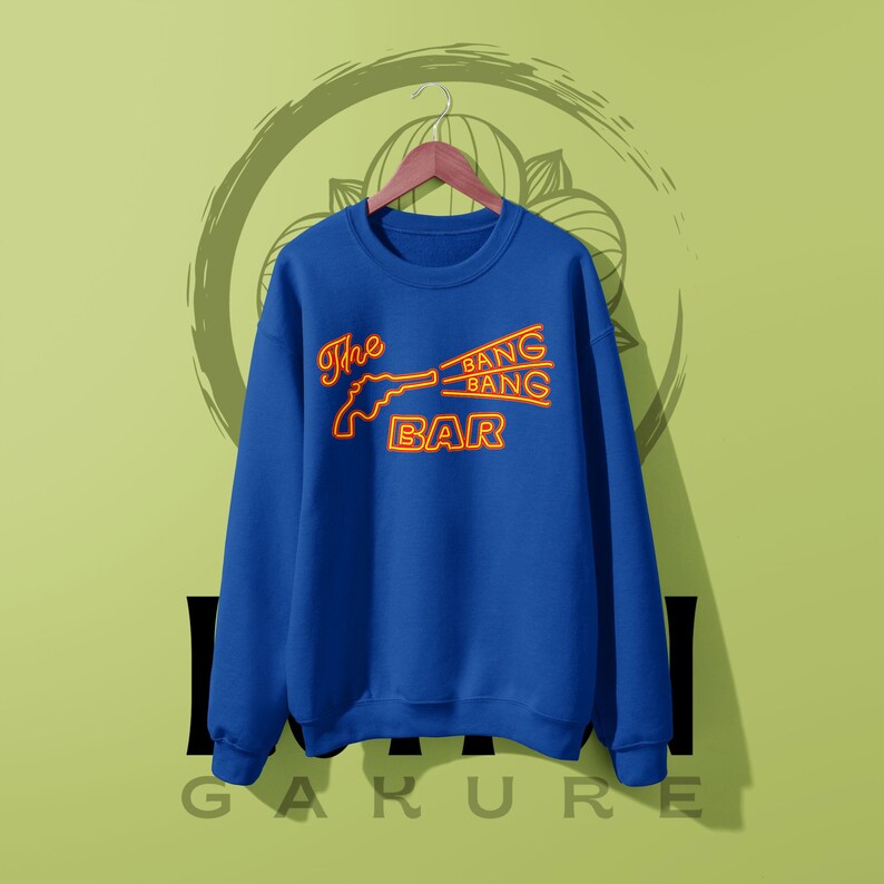 May include: A royal blue crewneck sweatshirt featuring a neon-style orange graphic on the chest. The design depicts a revolver silhouette with the text "The BANG BANG BAR" printed in a stylized, glowing orange font across the front of the garment.