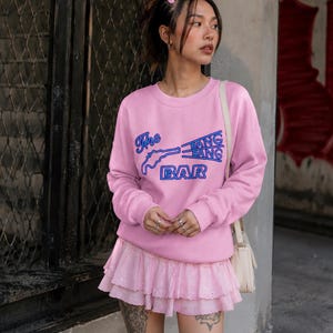 May include: A light pink crewneck sweatshirt featuring a blue graphic print of a pistol and the text The BANG BANG BAR. The sweatshirt has long sleeves with ribbed cuffs and a matching light pink tiered mini skirt with eyelet lace detailing.
