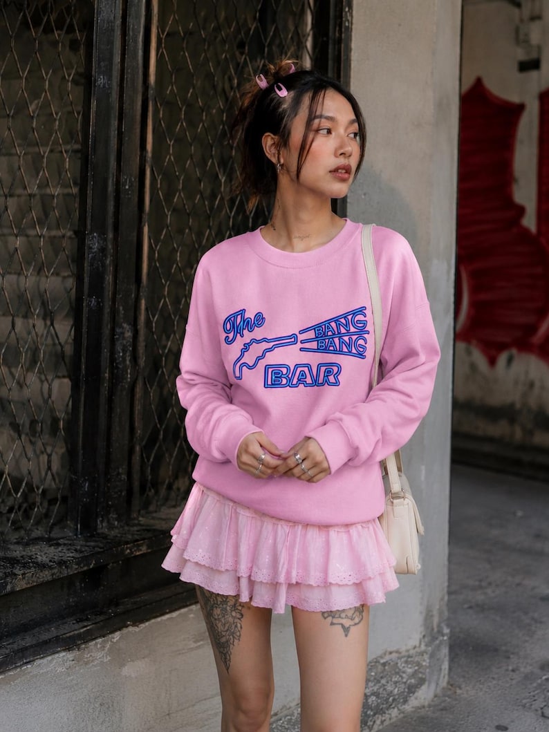 May include: A light pink crewneck sweatshirt featuring a blue graphic print of a pistol and the text The BANG BANG BAR. The sweatshirt has long sleeves with ribbed cuffs and a matching light pink tiered mini skirt with eyelet lace detailing.