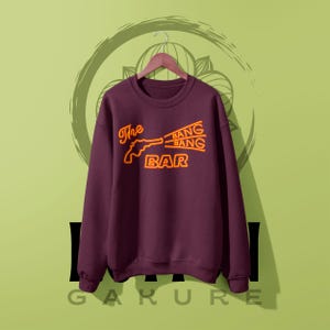 May include: A dark maroon crewneck sweatshirt featuring a neon-style graphic in orange and yellow. The design depicts a revolver silhouette with the text The BANG BANG BAR printed in a stylized font across the chest, hanging on a wooden hanger.