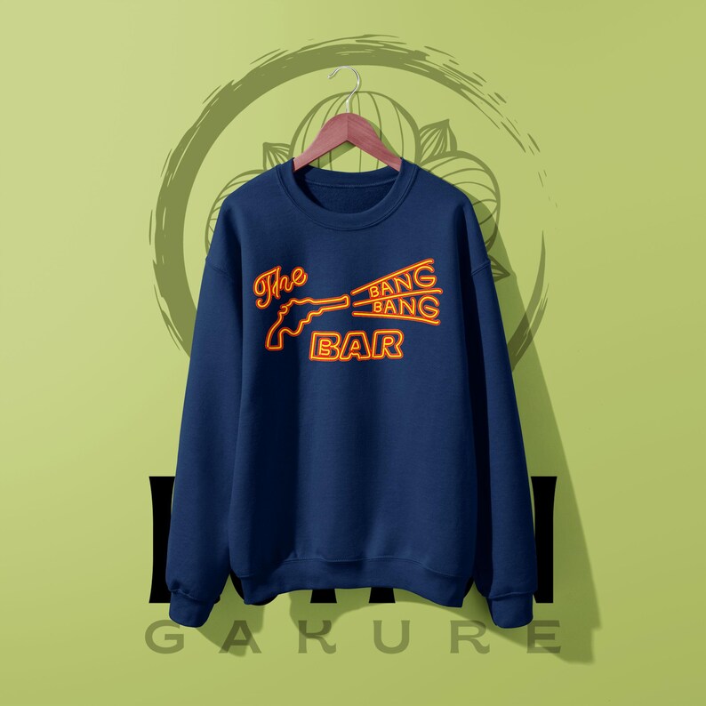 May include: A navy blue crewneck sweatshirt featuring a neon-style graphic in orange and yellow. The design depicts a revolver silhouette with the text The BANG BANG BAR, with the words BANG BANG emanating from the barrel in a stylized font.