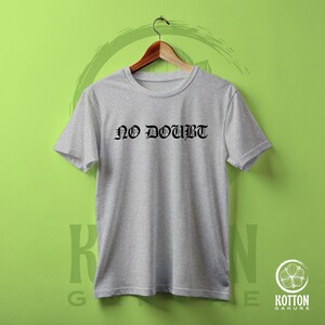 Puede incluir: Camiseta gris claro con la frase "NO DOUBT" en letras g&oacute;ticas negras. La camiseta est&aacute; colgada en una percha de madera sobre un fondo verde.