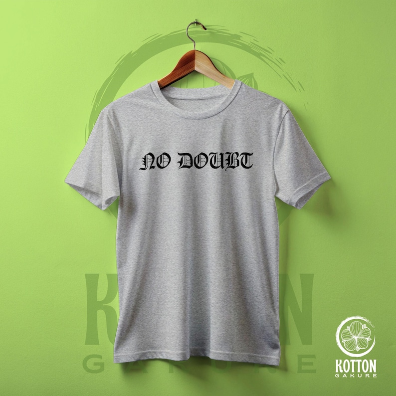 Puede incluir: Camiseta gris claro con la frase "NO DOUBT" en letras g&oacute;ticas negras. La camiseta est&aacute; colgada en una percha de madera sobre un fondo verde.