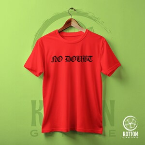 Puede incluir: Una camiseta roja brillante con la frase "NO DOUBT" impresa en letras g&oacute;ticas negras. La camiseta est&aacute; colgada en una percha de madera sobre un fondo verde. La camiseta es de cuello redondo.