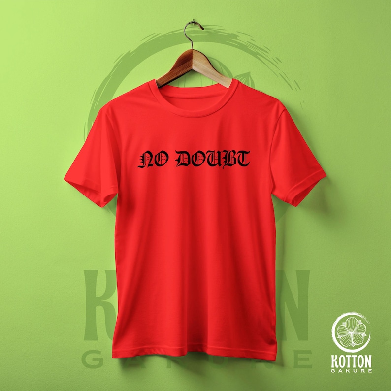 Puede incluir: Una camiseta roja brillante con la frase "NO DOUBT" impresa en letras g&oacute;ticas negras. La camiseta est&aacute; colgada en una percha de madera sobre un fondo verde. La camiseta es de cuello redondo.