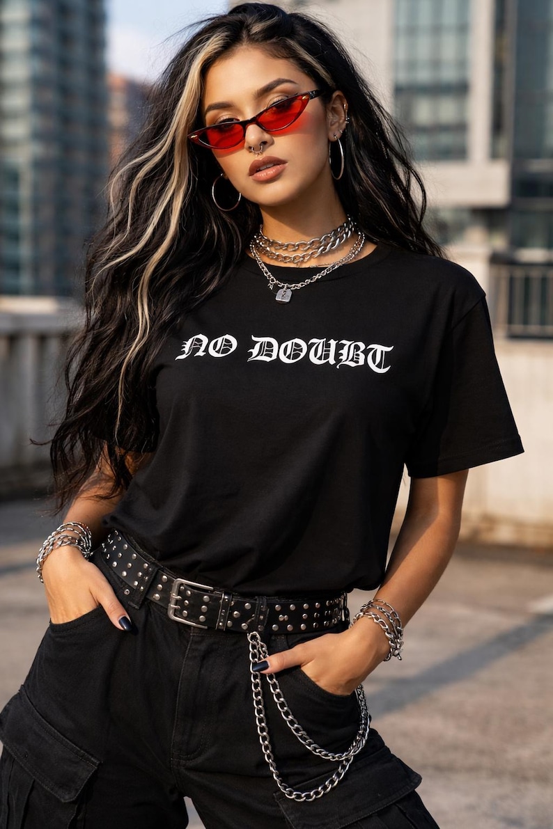 Puede incluir: Camiseta negra con el texto "NO DOUBT" en letras g&oacute;ticas blancas. La persona lleva un cintur&oacute;n con tachuelas, joyas de cadena plateada y gafas de sol rojas. El atuendo se completa con pantalones negros.