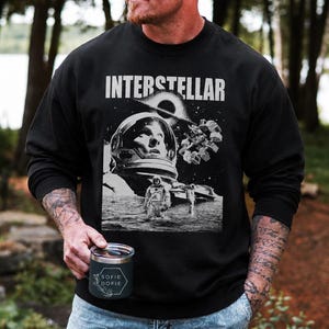 May include: A black long-sleeve sweatshirt featuring a white graphic print of the movie Interstellar. The design includes an astronaut helmet, a space station, and a black hole. The text INTERSTELLAR is printed in white capital letters at the top.