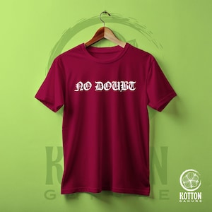 Puede incluir: Una camiseta granate con las palabras "NO DOUBT" en letras g&oacute;ticas blancas. La camiseta est&aacute; colgada de una percha de madera sobre un fondo verde. La camiseta es de cuello redondo y manga corta.