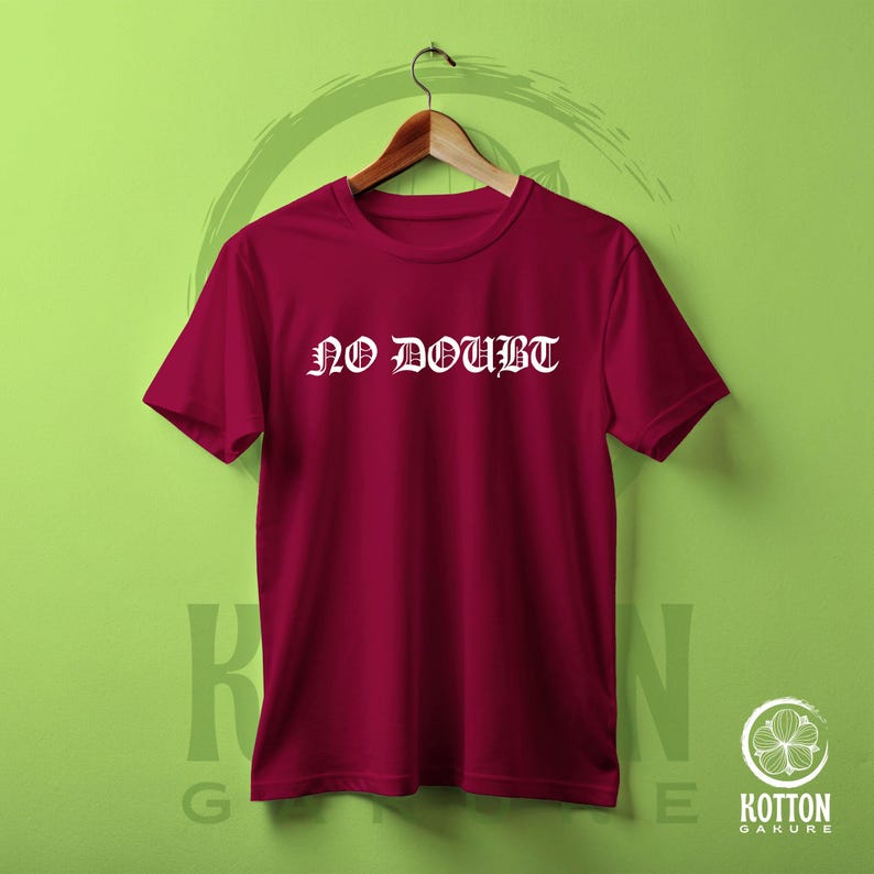 Puede incluir: Una camiseta granate con las palabras "NO DOUBT" en letras g&oacute;ticas blancas. La camiseta est&aacute; colgada de una percha de madera sobre un fondo verde. La camiseta es de cuello redondo y manga corta.