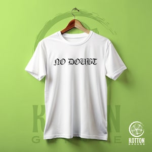 Puede incluir: Una camiseta blanca con la frase "NO DOUBT" impresa en negro con letras g&oacute;ticas. La camiseta est&aacute; colgada en una percha de madera sobre un fondo verde. Tiene mangas cortas y cuello redondo.