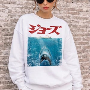 May include: A white long-sleeve crewneck sweatshirt featuring a graphic print of a shark swimming beneath a person in the ocean. Above the water, the Japanese text "ジョーズ" is printed in a distressed red font. The sweatshirt has a ribbed collar, cuffs, and hem.