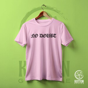 Puede incluir: Camiseta rosa claro con la frase "NO DOUBT" impresa en letras g&oacute;ticas negras. La camiseta est&aacute; colgada de una percha de madera sobre un fondo verde. La camiseta es de cuello redondo.