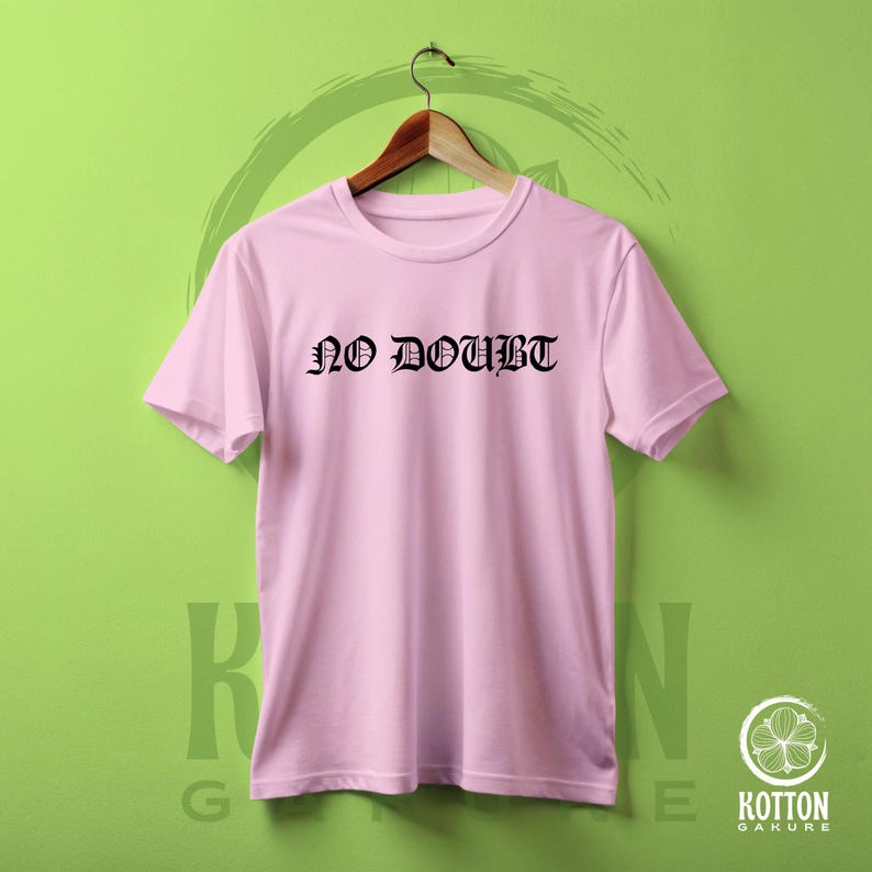 Puede incluir: Camiseta rosa claro con la frase "NO DOUBT" impresa en letras g&oacute;ticas negras. La camiseta est&aacute; colgada de una percha de madera sobre un fondo verde. La camiseta es de cuello redondo.