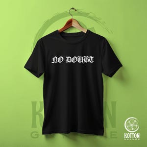 Puede incluir: Camiseta negra de cuello redondo con el texto blanco "NO DOUBT" en fuente g&oacute;tica. La camiseta est&aacute; colgada de una percha de madera sobre un fondo verde. Es una camiseta b&aacute;sica.