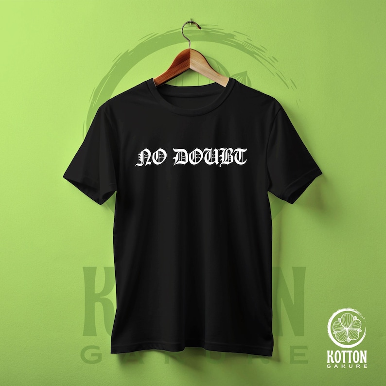 Puede incluir: Camiseta negra de cuello redondo con el texto blanco "NO DOUBT" en fuente g&oacute;tica. La camiseta est&aacute; colgada de una percha de madera sobre un fondo verde. Es una camiseta b&aacute;sica.