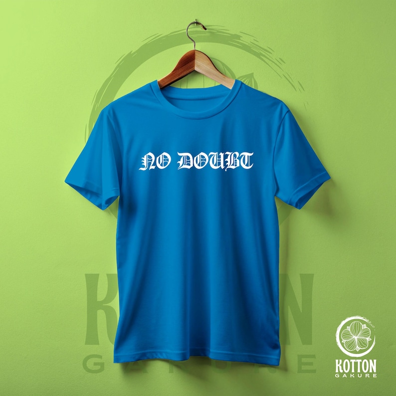 Puede incluir: Una camiseta azul con el texto blanco "NO DOUBT" en fuente g&oacute;tica. La camiseta est&aacute; colgada en una percha de madera sobre un fondo verde. La camiseta es de cuello redondo.