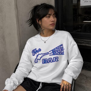 May include: A white long-sleeve crewneck sweatshirt featuring a blue graphic print of a revolver and the text The BANG BANG BAR. The design uses a bold, outlined font style with a neon-like blue color scheme across the center of the chest.