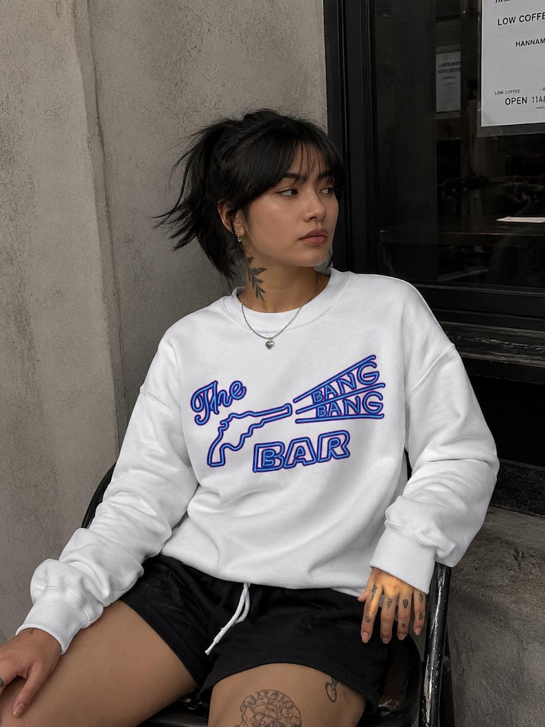 May include: A white long-sleeve crewneck sweatshirt featuring a blue graphic print of a revolver and the text The BANG BANG BAR. The design uses a bold, outlined font style with a neon-like blue color scheme across the center of the chest.