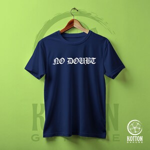 Puede incluir: Una camiseta azul marino con el texto blanco "NO DOUBT" en fuente g&oacute;tica. La camiseta est&aacute; colgada de una percha de madera sobre un fondo verde. La camiseta es de cuello redondo.