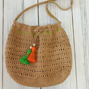 May include: A woven brown shoulder bag with a drawstring closure. The bag features a decorative orange and green embroidered trim along the top and a pair of matching tassels. The strap is also woven.