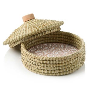 May include: A round woven basket with a lid, featuring a natural straw color and a wooden knob on the lid. The basket has a pink and white ceramic plate inside.