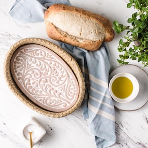 May include: A woven oval serving dish with a pink and white floral design. The dish is on a white marble surface with a loaf of bread and a bowl of olive oil.