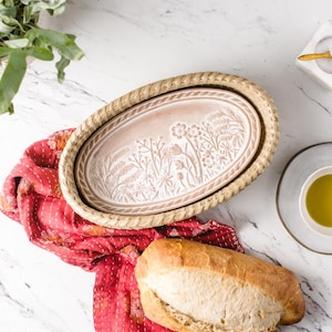 Może przedstawiać: Ceramiczna forma do pieczenia chleba z kwiatowym wzorem, osadzona w plecionym koszyku. Forma jest jasnobrązowa z białymi kwiatowymi detalami. Bochenek chleba leży na czerwonej tkaninie.