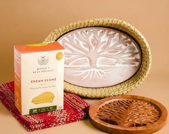 May include: A cream scone mix box with the text "Women's Bean Project" and a scone illustration sits on a red patterned cloth. A large oval woven tray and a wooden carved tray with a tree design are also present.