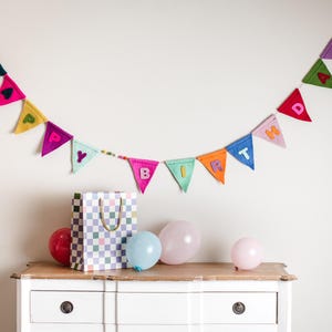 Può includere: Uno striscione di feltro colorato con le parole "Happy Birthday" appeso a un muro bianco. Un cassettone bianco con un cassetto è in primo piano. Ci sono anche tre palloncini rosa e una borsa regalo con un motivo a scacchi.
