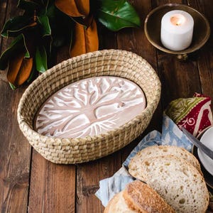 May include: A woven oval basket with a ceramic tree of life design inside. The basket is made of natural materials and has a rustic look. There are three loaves of bread on a blue and white patterned cloth in the foreground.