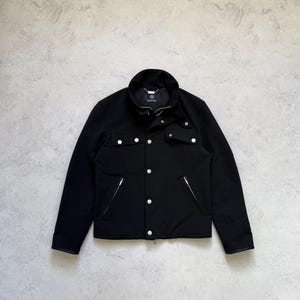 May include: A black jacket featuring a high collar, a front button closure with silver-toned hardware, two chest pockets with button flaps, and two lower slanted pockets with zipper closures. The interior label displays the Versace brand name.