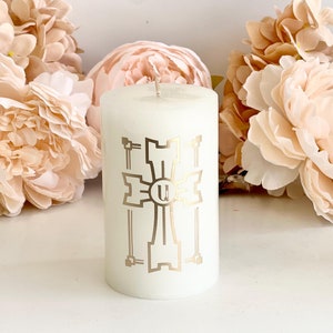 Puede incluir: Vela blanca en forma de pilar con un diseño de cruz dorada y la letra "U" en el centro. La vela está rodeada de flores rosas.