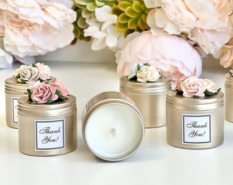 Könnte beinhalten: Goldfarbene Kerzen in Dosen mit Blumenschmuck und einem "Thank You!" Etikett. Jede Kerze ist mit kleinen, zarten Blüten in Rosa und Creme verziert. Der Hintergrund zeigt sanfte, rosafarbene Blumenarrangements.