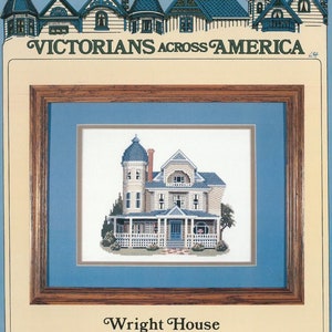 May include: A cross-stitch pattern of a Victorian house called the Wright House in Eureka Springs, Arkansas. The house is white with a blue roof and a porch with white railings. The pattern is framed in a brown wooden frame with a blue mount.