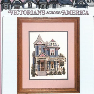 May include: A framed cross-stitch of a Victorian house in blue, pink, and white, with a wooden frame. The text "VICTORIANS ACROSS AMERICA" is at the top, and "Druid Circle Atlanta, Georgia" is at the bottom.