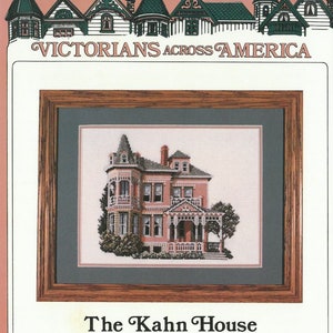 May include: A cross-stitch pattern of a pink Victorian house with a porch and a green tree in front. The pattern is framed in a brown wood frame. The text "The Kahn House Ogden, Utah" is below the frame.