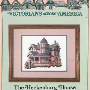 May include: A cross-stitch pattern of a Victorian house, called "The Heckenburg House", located in Lakewood, Ohio. The house is pink with white trim and a grey roof. The pattern is framed in a brown frame.