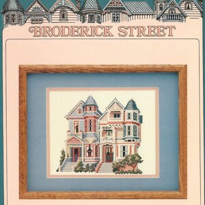 May include: A framed cross-stitch design of two colourful Victorian houses with a blue sky background. The title "BRODERICK STREET" is above the frame.