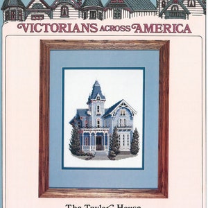 May include: A framed cross-stitch depicting a Victorian house in shades of blue and white. The artwork is presented with a light blue mount and a wooden frame. The text "VICTORIANS ACROSS AMERICA" is at the top, and "The Taylor House, Earlville, New York" is at the bottom.