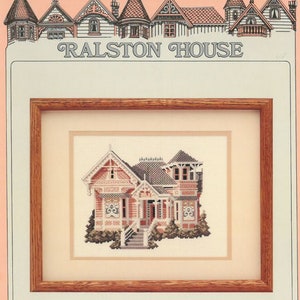 May include: A cross-stitch pattern of a pink Victorian house with a white picket fence and a green lawn. The pattern is framed in a brown wooden frame. The title "Ralston House" is printed above the frame.