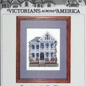 May include: A framed cross-stitch pattern of a Victorian house with a white facade and blue trim. The house has a wraparound porch with white railings and a black gate. The house is surrounded by green bushes and trees. The text "Victorians Across America" is at the top of the image. The text "Reymenschoffer House Galveston, Texas" is at the bottom of the image.