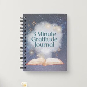 Pode incluir: Um diário com espiral com um design de céu estrelado. A capa apresenta o texto "3 Minute Gratitude Journal" em verde-azulado, com uma ilustração de um livro aberto. O diário está sobre uma superfície branca.